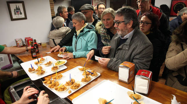 Varias personas esperan su turno para recoger los pinchos de verdura dispuestos en la barra de la peña Athletic de Tudela.
