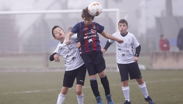 Un jugador del Mendigoiti, con camiseta azulgrana, trata de rematar de cabeza ante la presión de un futbolista del Sagrado Corazón, de blanco. Detrás, un compañero sigue la jugada.