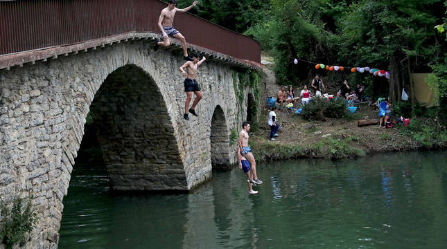 Varios jóvenes se lanzan al Arga desde el puente de Irotz, mientras otro grupo festeja en la orilla.