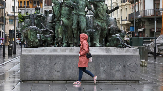 Una joven pasa ante el monumento al encierro de Pamplona cubierta con capucha y mascarilla.
