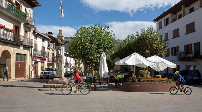 Foto de dos menores pasando en bicicleta por delante de la 'picota' de Lakuntza, vestigio de castigo de fechorías.